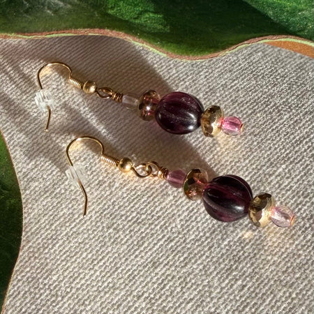 A pair of gold-toned earrings with golden, pink and clear, and deep amethyst-colored beads lies on ivory fabric, with green leaves to the top and side. The golden beads are faceted; the pink and clear ones, smooth; and the purple ones, fluted.