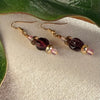 A pair of earrings with golden, pink, and deep amethyst-purple beads lies on ivory fabric, with green leaves to the top and side.