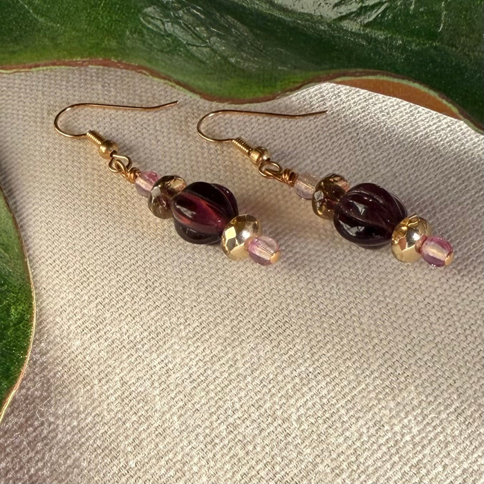 A pair of earrings with golden, pink, and deep amethyst-purple beads lies on ivory fabric, with green leaves to the top and side.