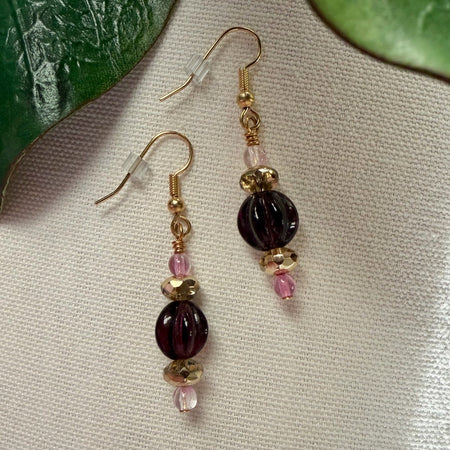 A pair of gold-toned earrings with purple, pink, and golden beads lies on ivory fabric, with green leaves in the top corners of the photo.