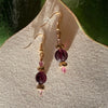 A pair of gold-toned earrings featuring pink, purple, and golden beads lies on ivory fabric, with green leaves in the top corners of the photo. The earrings cast shadows on the fabric.