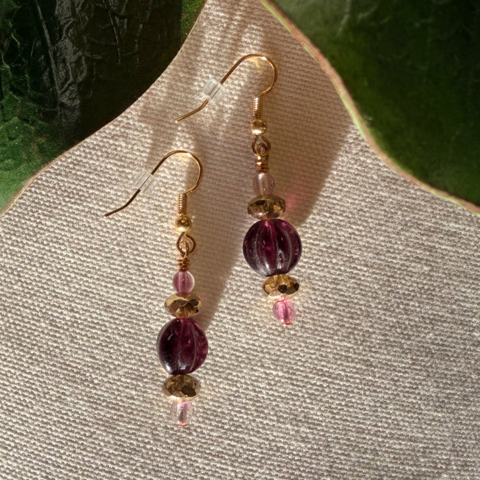 A pair of gold-toned earrings featuring pink, purple, and golden beads lies on ivory fabric, with green leaves in the top corners of the photo. The earrings cast shadows on the fabric.