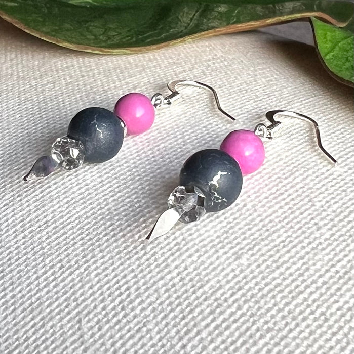 A pair of earrings with orchid-pink, silver, and silver-crackled gray beads lies diagonally on a textured ivory surface, with green leaves in the upper part of the photo.
