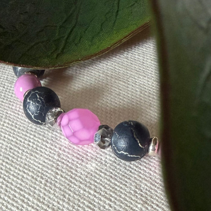 Part of a strand of beads lies on textured ivory fabric, with green leaves at the top and side. The beads are silver, dark gray with a silver crackle effect, and orchid pink; one of the pink beads features a snakeskin pattern.