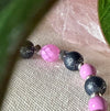 Part of a strand of beads lies on textured ivory fabric. The beads are silver-toned, orchid pink, and dark gray. One pink bead has a snakeskin pattern, and the gray beads feature a silver crackle effect. To the sides of the photo are green leaves; near the top is part of a light-pink, translucent object.
