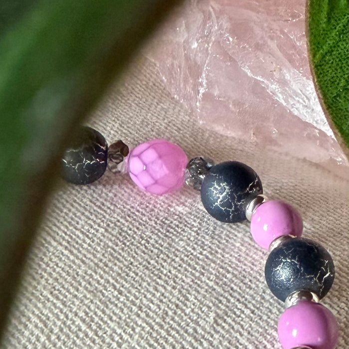 Part of a strand of beads lies on textured ivory fabric. The beads are silver-toned, orchid pink, and dark gray. One pink bead has a snakeskin pattern, and the gray beads feature a silver crackle effect. To the sides of the photo are green leaves; near the top is part of a light-pink, translucent object.
