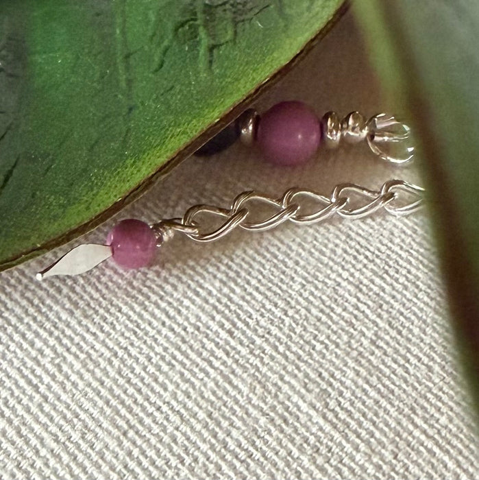 A few links of silver-toned chain, ending in a silver-toned spearhead-shaped dangle accented with an orchid-pink bead, lies on textured ivory fabric. In the background a larger pink bead and silver ones are visible. In the top corners of the photo are green leaves.