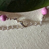 A silver-toned lobster-claw clasp and a length of chain lie on textured ivory fabric. To each side is an orchid-pink bead, and a green leaf appears in the top portion of the photo.