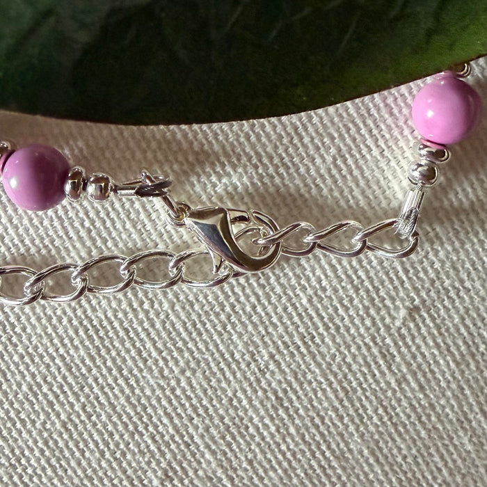 A silver-toned lobster-claw clasp and a length of chain lie on textured ivory fabric. To each side is an orchid-pink bead, and a green leaf appears in the top portion of the photo.