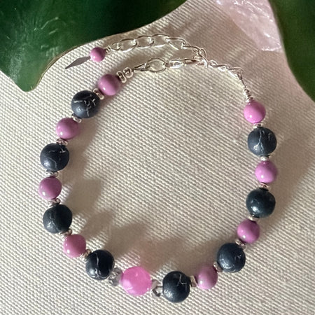 A silver-toned bracelet with orchid-pink and dark-gray beads lies on textured ivory fabric. The gray beads have a silver crackle effect. At the top of the photo are two green leaves with part of a light-pink translucent object between them.