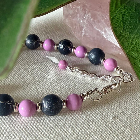 Part of a strand of silver-toned, orchid-pink, and dark-gray beads with a silver-toned chain and findings lies on textured ivory fabric. The gray beads feature a silver crackle effect. At the top of the photo are green leaves and a light-pink, translucent object.