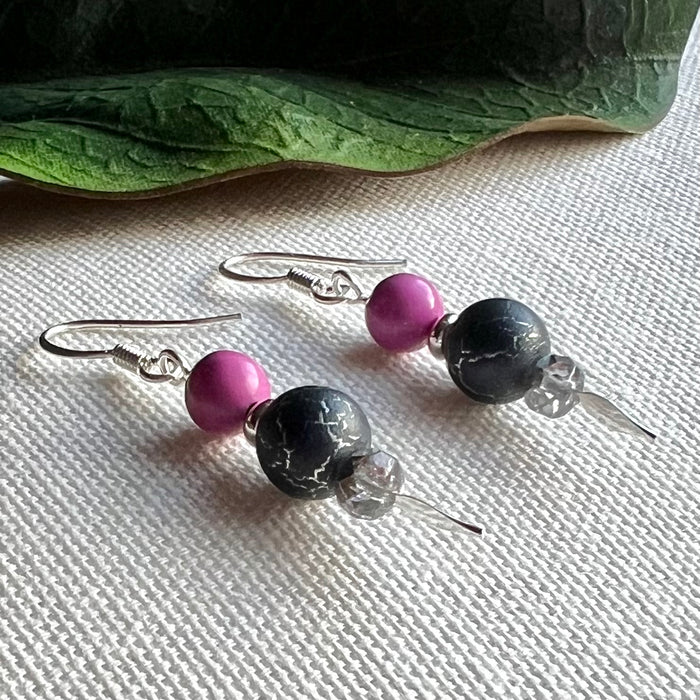 A pair of earrings featuring orchid-pink, silver, and silver-crackled gray beads lies diagonally on a textured ivory background, with a green leaf across the upper part of the photo.