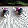 A pair of earrings with orchid-pink, silver, and silver-crackled gray beads lies on a textured ivory surface, with a green leaf in the background. The camera angle foregrounds the bottom tips of the earrings.