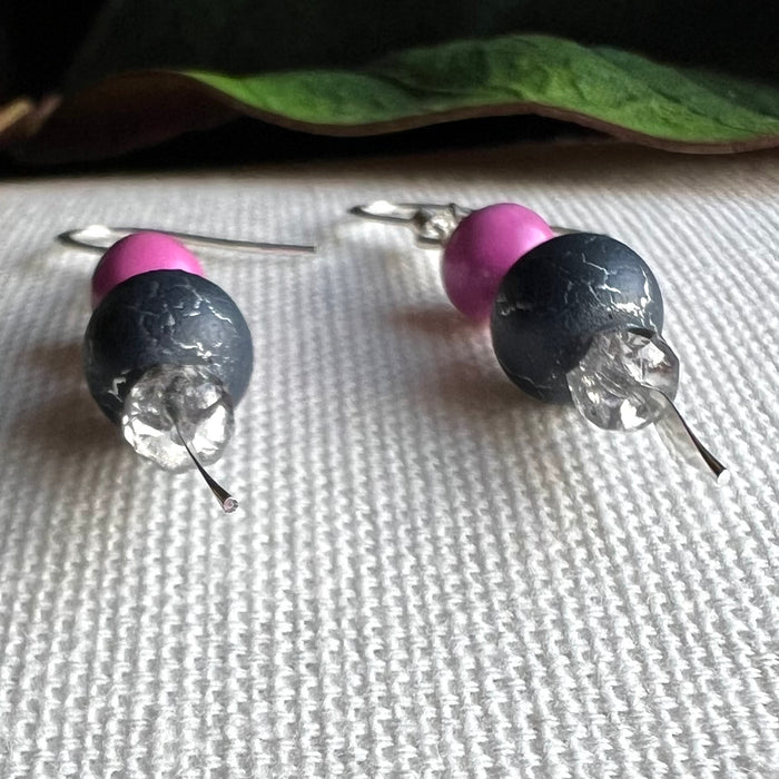 A pair of earrings with orchid-pink, silver, and silver-crackled gray beads lies on a textured ivory surface, with a green leaf in the background. The camera angle foregrounds the bottom tips of the earrings.