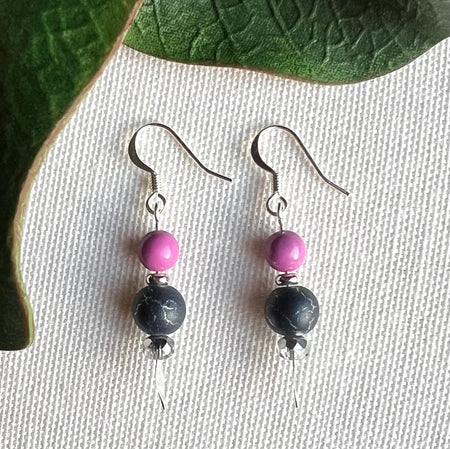 A pair of earrings featuring orchid-pink, silver, and silver-crackled gray beads lies on a textured ivory background, framed on two sides by green leaves.