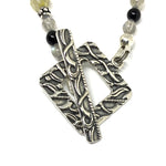 A square antiqued-silver toggle clasp attached to a strand of gray, black, silver, and yellow beads lies on a white background.