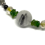 A strand of beads lies on a white background. Near the center is a large faceted bead, cloudy white with black needle shapes inside; that bead is bookended by smaller green stone chips, then yellow-golden beads, and at one end a black and a silver bead.