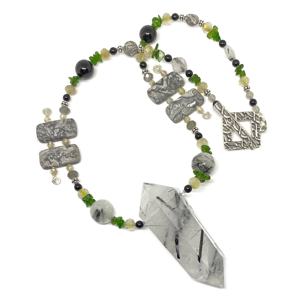 A black, white, gray, green, yellow, and silver beaded necklace with a white, gray, and black tower-shaped stone pendant lies on a white background, curved so that the whole strand is visible.