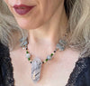 A woman wearing burgundy lip color and a lace-trimmed black top models a white, gray, black, yellow, green, and silver necklace with a large black and white tower-shaped pendant.