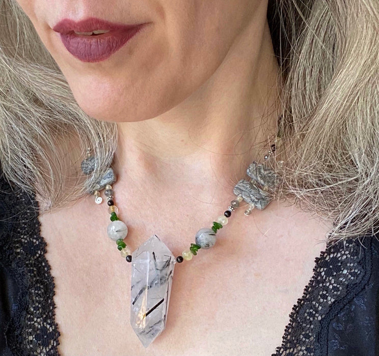 A woman wearing burgundy lip color and a lace-trimmed black top models a white, gray, black, yellow, green, and silver necklace with a large black and white tower-shaped pendant.