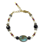A beaded bracelet lies on a white background. The gray focal bead with flashes of blue is toward the bottom of the photo, and the golden extender chain toward the top. The colors of the beads include black, gray, white, red, and golden.