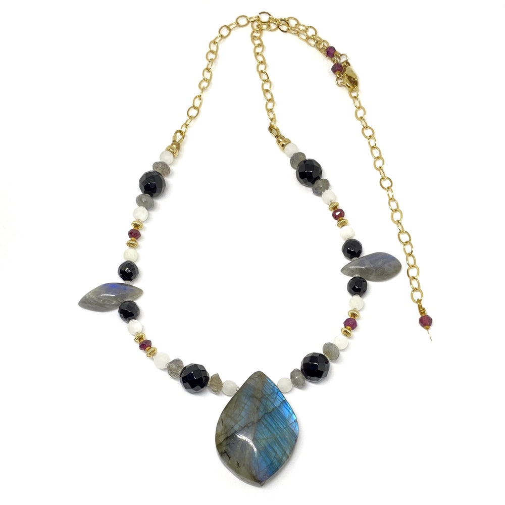 A pendant necklace featuring one large and two smaller gray gemstone focals lies on a white background. Part of the necklace is beaded with gray, black, white, red, and golden beads, and part of it consists of golden chain and an extender.