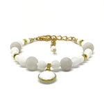 A bracelet with white and pale-gray beads, gold-toned accents, and a white and gold-toned charm lies on a white background. 