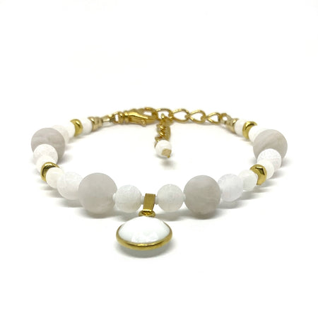 A bracelet with white and pale-gray beads, gold-toned accents, and a white and gold-toned charm lies on a white background. 