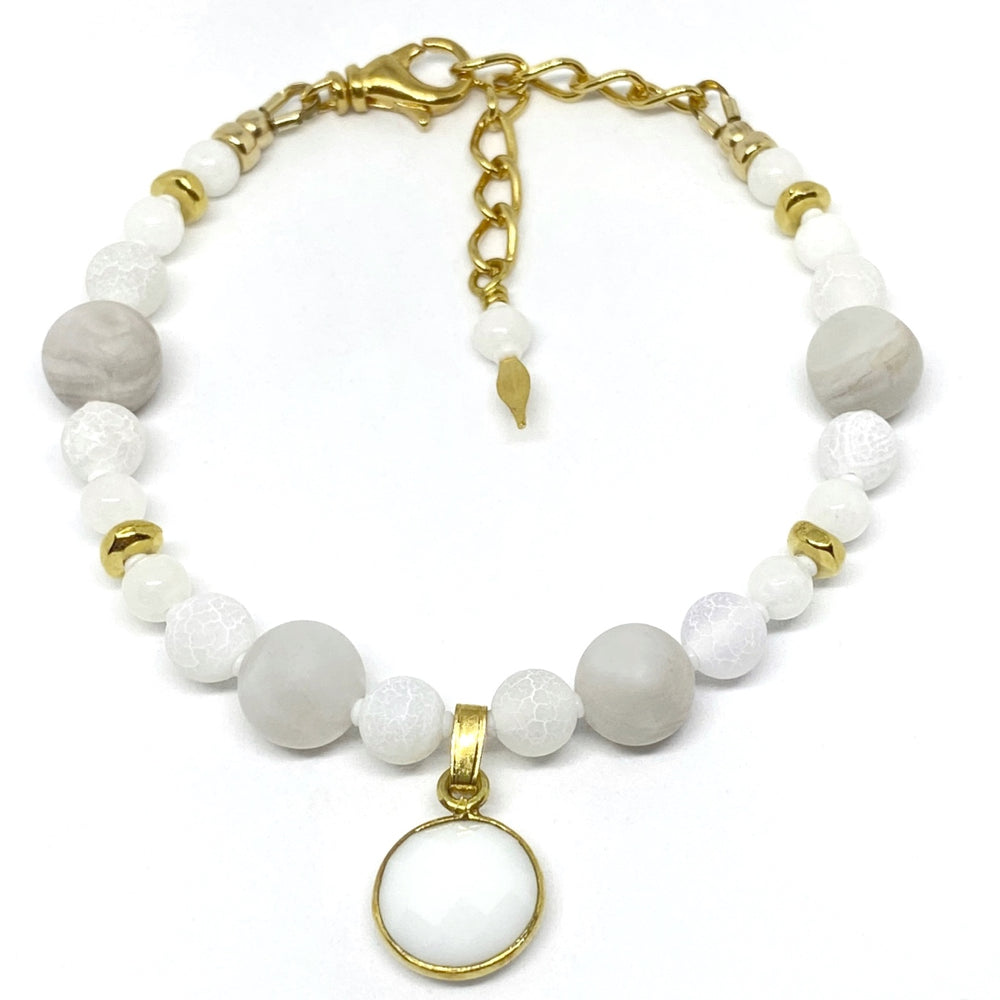 A bracelet with white and pale-gray beads and gold-toned accents lies on a white background. The bracelet features a circular, white and gold-toned charm and a gold-toned extender chain with a white bead and a spear-tipped headpin on the dangle.