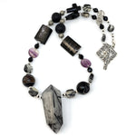On a white background lies a beaded necklace featuring a square toggle clasp, beads in shades of black, purple, and silver, and a large, tower-shaped pendant of gray stone with black needles formed inside it.