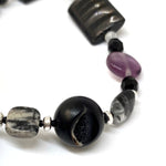 Part of a beaded strand of jewelry is visible. In the center foreground is a black bead with an opening inside which druzy can be seen. The other beads are silver, black, gray, and purple in color.