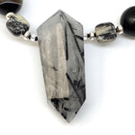 A pointed gray stone pendant with black needles formed inside lies on a white background. Beads are visible on a strand to either side of the pendant.