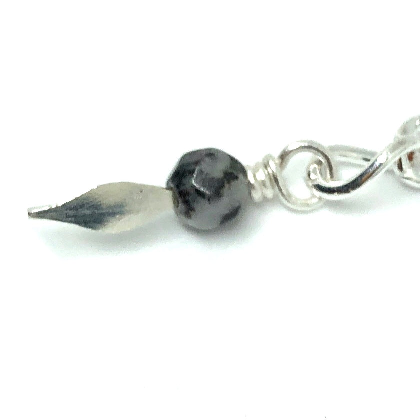 A beaded dangle sits in the center of the photo on a white background. The bead is a small, faceted black and white round. It is attached to a link of chain by a spear-shaped paddle pin. Both the pin and the chain are silver-toned.