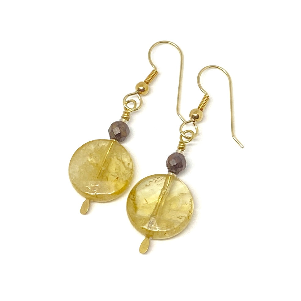 On a white background lies a pair of earrings. Each features a coin-shaped bead in a deep golden-yellow, topped by a small faceted wood bead and a golden earwire.