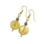 On a white background lies a pair of earrings. Each features a coin-shaped bead in a deep golden-yellow, topped by a small faceted wood bead and a golden earwire.