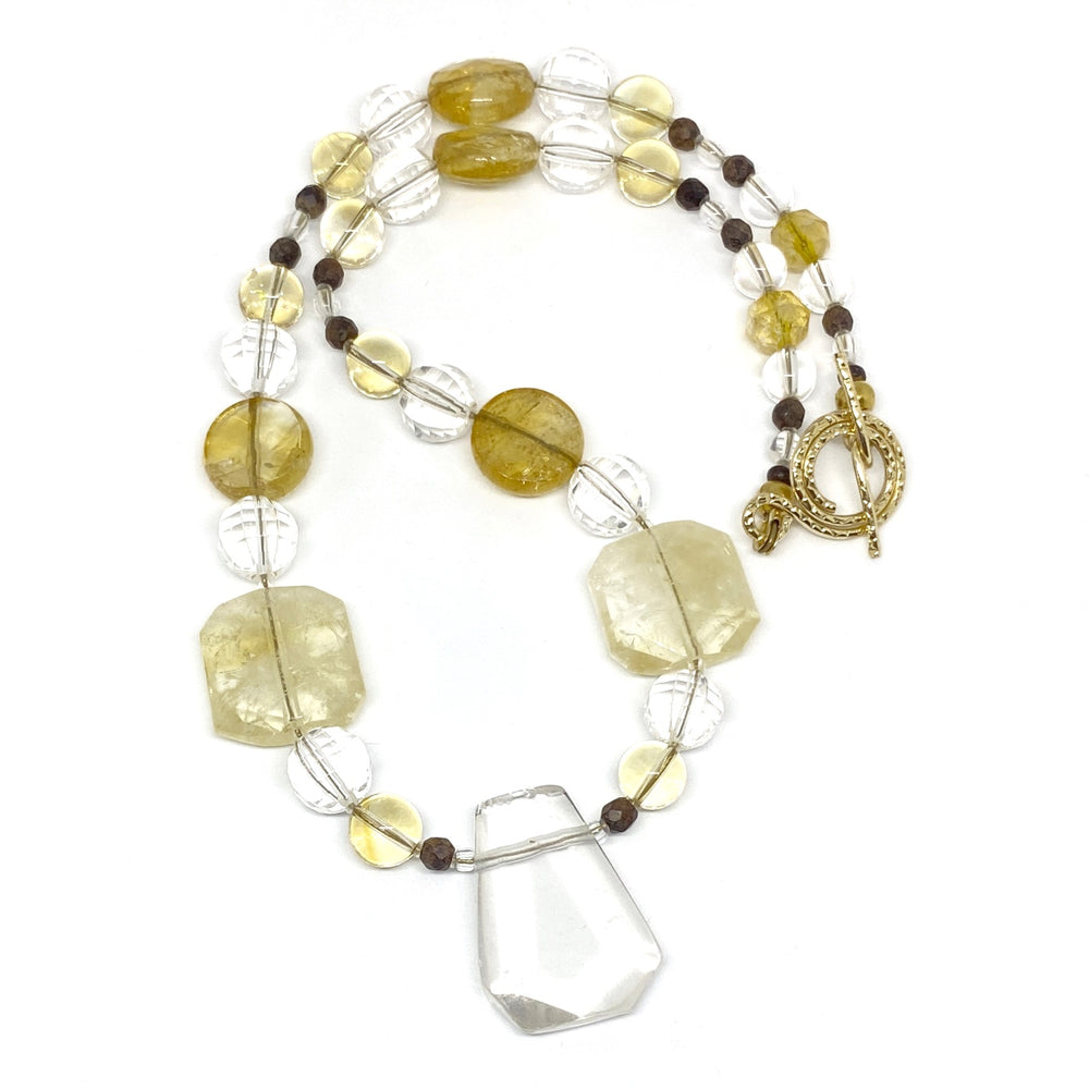 On a white background lies a beaded necklace with a transparent focal bead. The other beads range from transparent to light yellow to deep golden tones and are accented by small wood beads.. The clasp is a golden textured toggle.